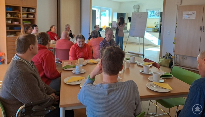 Mehrere Teilnehmer sitzen an einem gro&szlig;en Tisch bei Kaffee und Kuchen und verfolgen eine Erkl&auml;rung vor einer Tafel.