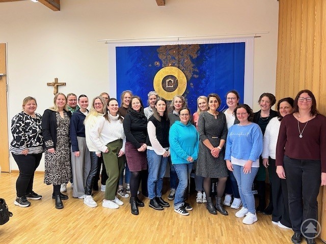 Diese Caritas-Kita-Leiterinnen sind nach den Zertifikats-Kursen persönlich und fachlich qualifiziert. Mehrere Frauen stehen in einem Gruppenfoto in einem Raum vor einem großen blauen Wandbild mit goldener Gestaltung und blicken in die Kamera.