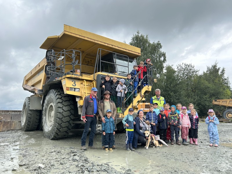 Kinder und Begleitpersonen des Schönberger Ferienprogramms vor einem beeindruckenden Muldenkipper im Steinbruch Thiele. Eine Gruppe von Kindern und Erwachsenen steht vor einem riesigen gelben Muldenkipper im Steinbruch, einige Kinder sitzen auf der Treppe des Fahrzeugs.