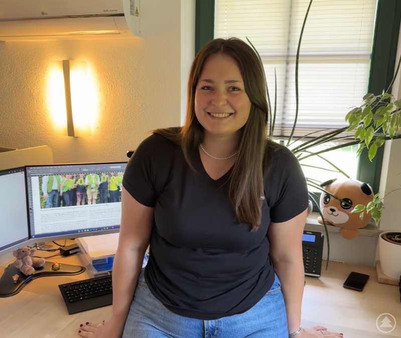 Amelie Fuchs - unsere Grafikmaus Auf dem Bild sitzt eine junge Frau lächelnd auf einem Schreibtisch in einem modernen Büro. Sie trägt ein schwarzes T-Shirt und eine Jeans. Hinter ihr stehen zwei Monitore, auf einem ist ein Text mit einem Bild von Personen in neon grünen T-Shirts geöffnet. Auf dem Schreibtisch liegen eine Tastatur, ein Telefon, ein Stofftier und einige Büroutensilien. Rechts am Fenster ist eine grüne Pflanze zu sehen, daneben ein rundes Kissen oder Stofftier mit einem Tiergesicht. Im Hintergrund fällt durch ein Fenster Tageslicht herein.