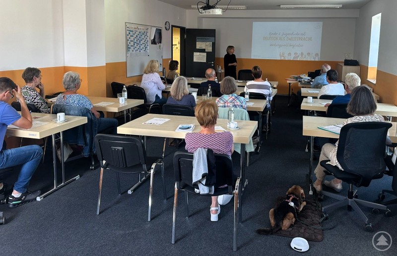 Wichtige Grundlagen und bewährte Methoden zur Unterstützung von Kindern mit Deutsch als Zweitsprache im schulischen Umfeld lernten Sprachpatinnen und Sprachpaten und solche, die es vielleicht noch werden wollen, im Rahmen einer Schulung kennen. Mehrere Personen sitzen in einem Seminarraum an Tischen und hören einer Referentin zu, die vorne vor einer Leinwand steht. Auf der Leinwand ist eine Präsentation mit dem Thema „Deutsch als Zweitsprache“ zu sehen. Im Vordergrund liegt ein Hund auf einer Decke am Boden.