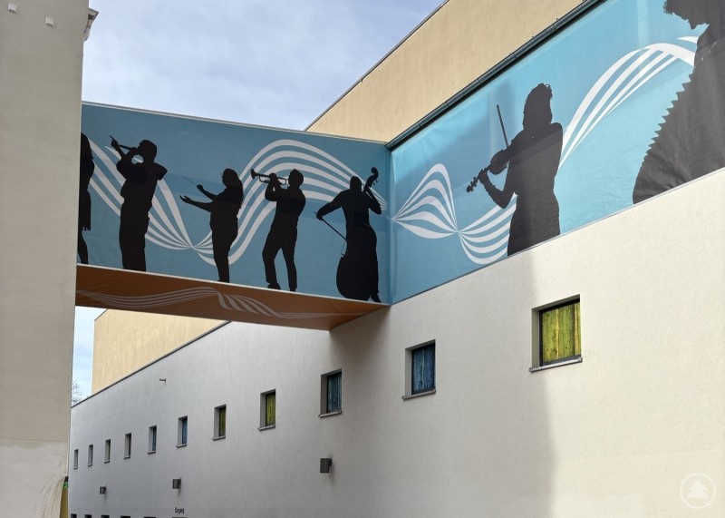 Der Verbindungssteg mit musikalischen Silhouetten führt zum zukünftigen Ausstellungsbereich des TonYversums. Ein schmaler Durchgang zwischen zwei Gebäuden, darüber ein bemalter Steg mit Musikern in Silhouettenoptik.