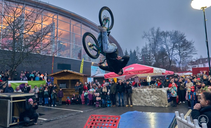 Ein Street-Trial-Biker zeigt einen spektakul&auml;ren R&uuml;ckw&auml;rtssalto mit dem Fahrrad vor einer gro&szlig;en Menschenmenge. Zuschauer stehen dicht gedr&auml;ngt rund um die Aktionsfl&auml;che.