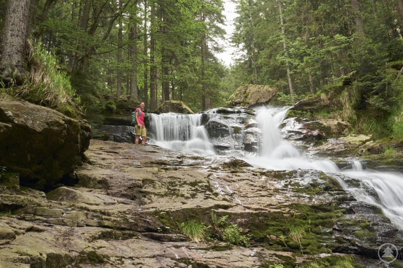 Eine Person steht auf Felsen neben einer breiten Wasserfall-Kaskade im Wald. 