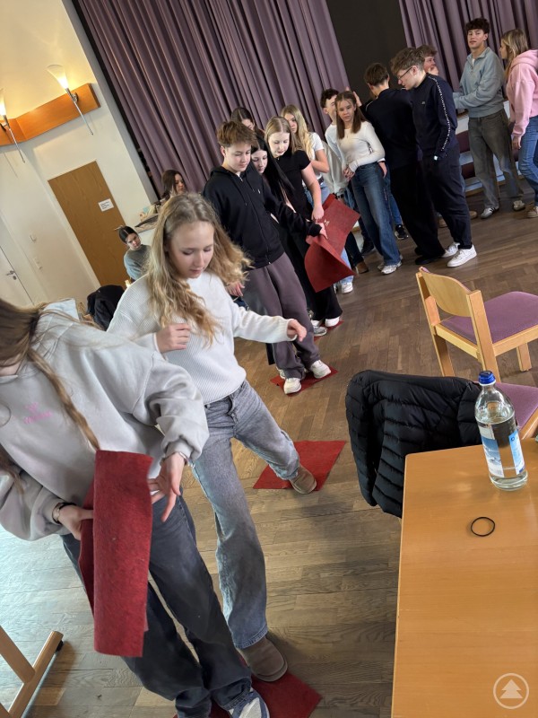 Sch&uuml;lerinnen und Sch&uuml;ler stehen im Kreis in einem gro&szlig;en Raum und halten Schn&uuml;re, die an einer Holzscheibe befestigt sind. In der Mitte versuchen sie gemeinsam, Holzkl&ouml;tze zu stapeln.