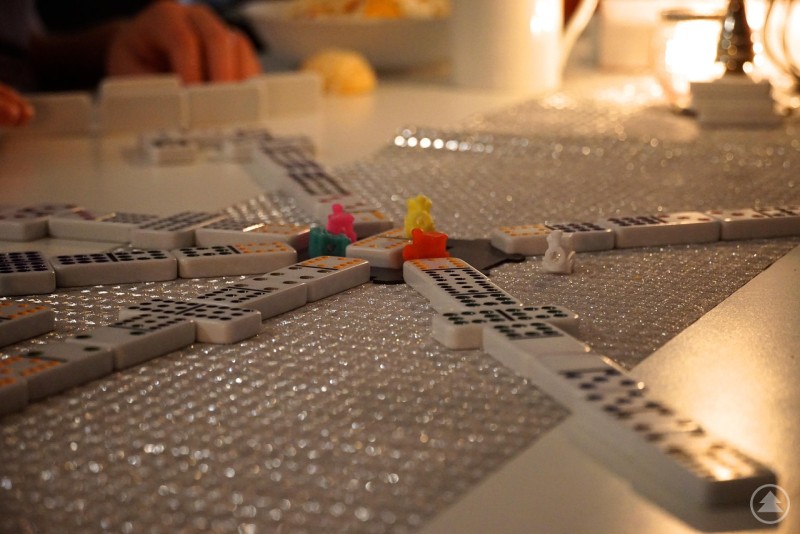 Das Bild zeigt ein geselliges Spiel mit Domino-Steinen auf einem glitzernden Tischläufer. Es handelt sich vermutlich um das Spiel „Mexican Train Dominoes“, erkennbar an den kleinen bunten Spielsteinen in Zugform, die auf verschiedenen Domino-Ketten stehen. Im Hintergrund sieht man Hände von Mitspielern, eine Schale mit Snacks und eine gemütliche Beleuchtung mit Kerze – die Szene wirkt warm und einladend.