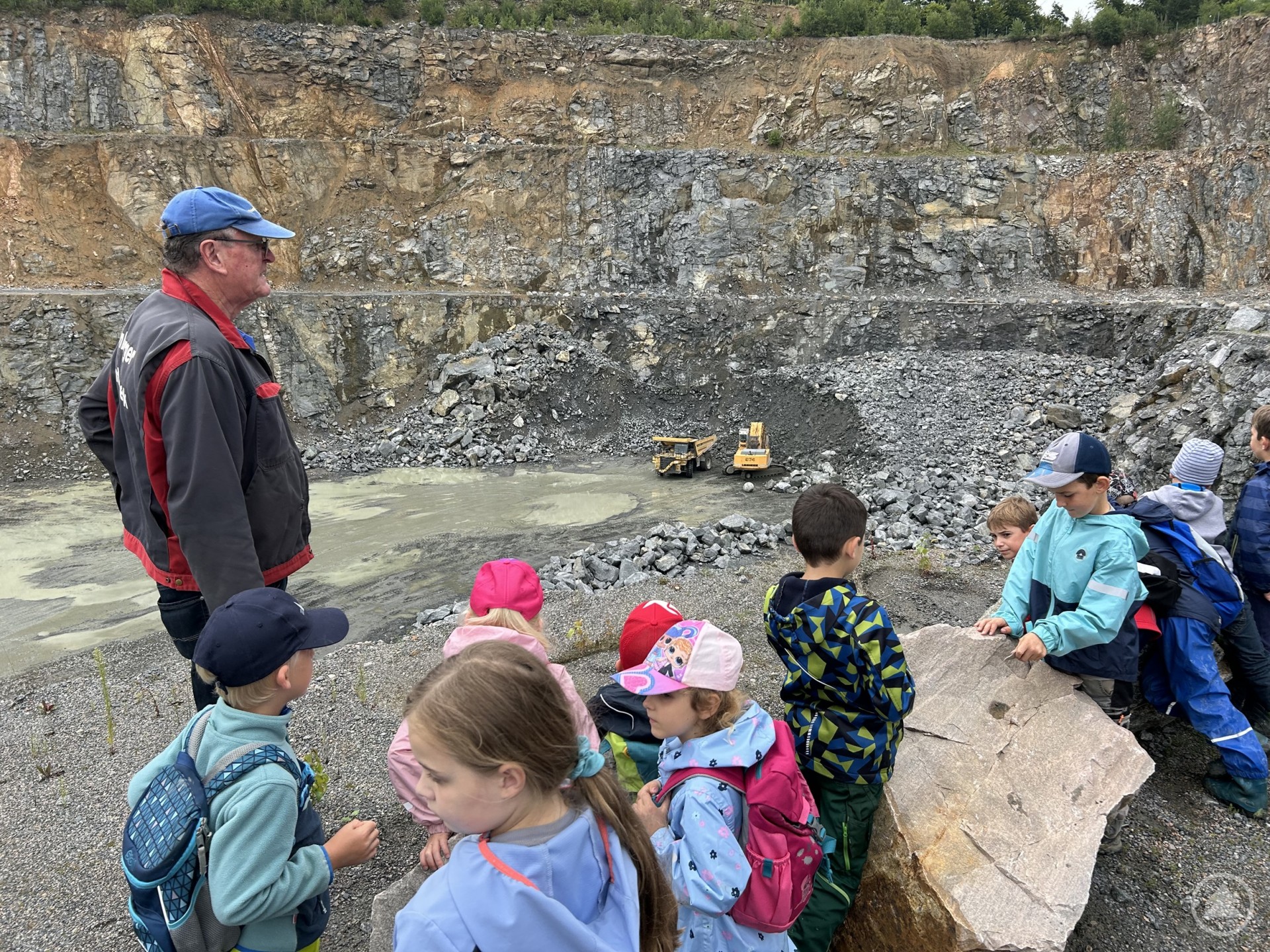 Kinder mit Rucksäcken und Mützen stehen am Rand eines Steinbruchs und blicken auf die großen Maschinen im Abbaugebiet.