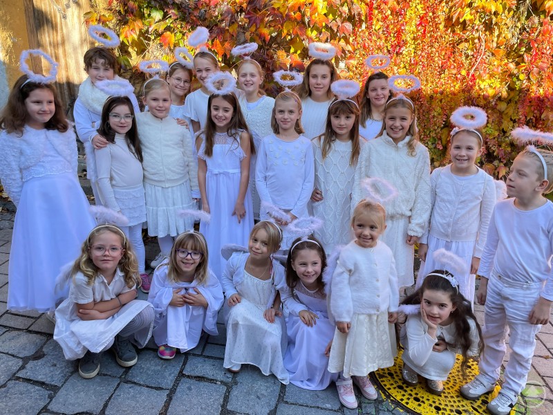 Die Kinderfreunde Schärding freuen sich auf „Kinder, Keks & Kabarett“ im Schärdinger Kubinsaal. Eine Gruppe von Kindern steht fröhlich beisammen, alle tragen weiße Engelskostüme mit Heiligenschein. Im Hintergrund ist eine herbstlich bunt bewachsene Mauer zu sehen.