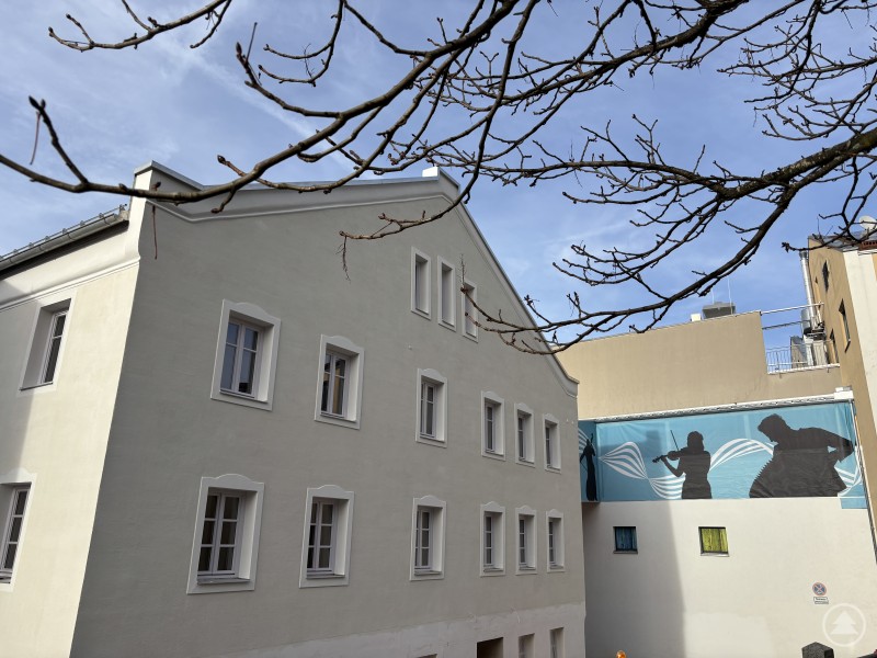 Ein frisch renoviertes Gebäude mit heller Fassade steht im Vordergrund, im Hintergrund ist ein darüber verlaufender Kunststeg mit Musikanten-Silhouetten zu sehen.