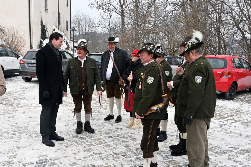 Austausch und Geselligkeit nach dem traditionellen Neujahrsanschießen am Schloss Wolfstein. Landrat Sebastian Gruber spricht im verschneiten Schlosshof mit einer Gruppe Böllerschützen in Tracht. Im Hintergrund stehen mehrere Autos und Bäume.