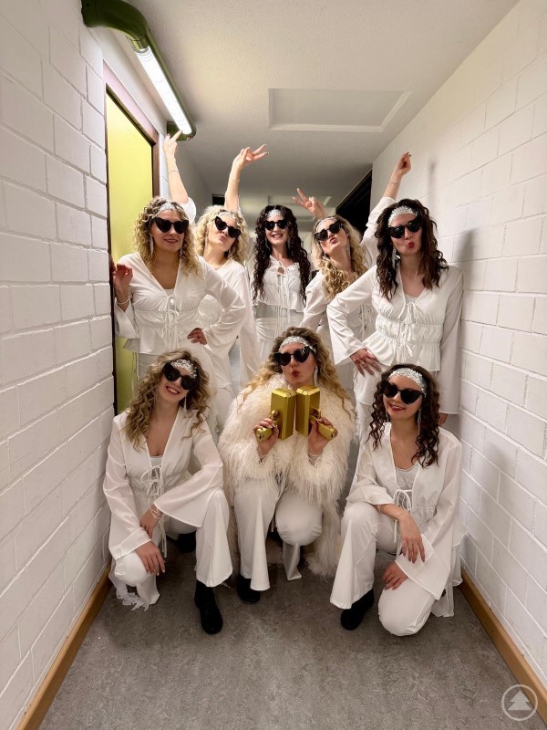 Backstage Stimmung vor dem Auftritt: Teamgeist wird bei der Garde Grafenau großgeschrieben. Die Tänzerinnen stehen lachend in einem schmalen Gang und präsentieren ihre weißen Kostüme mit Sonnenbrillen.