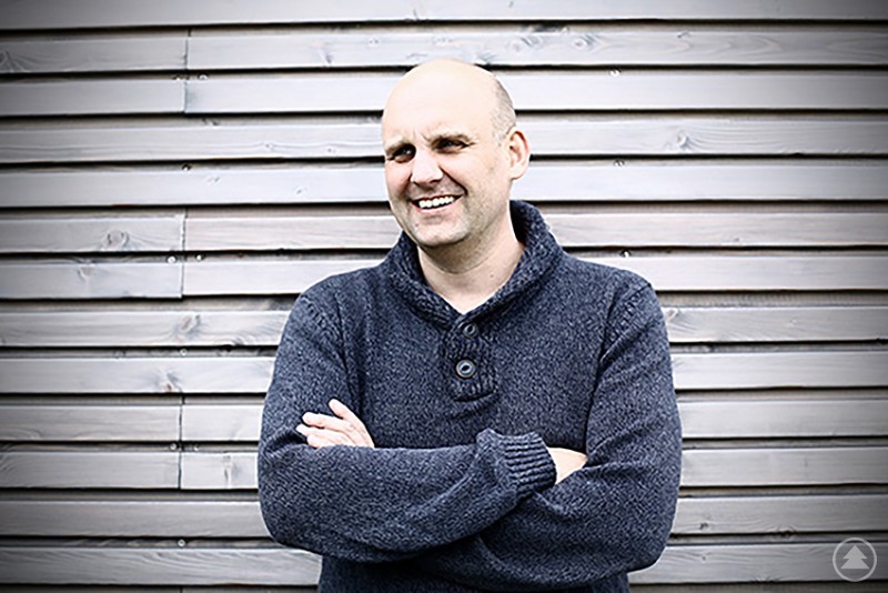 Kabarettist Christoph Maul gastiert in der Stadtbibliothek Deggendorf. Ein lächelnder Mann mit verschränkten Armen steht vor einer Holzwand und blickt freundlich in die Kamera.