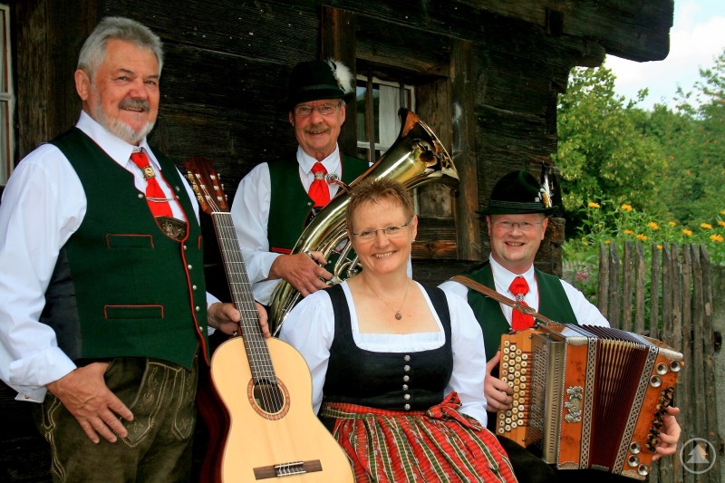 „d’Waidler Musi“ feiert ihr 20-jähriges Jubiläum mit einem Weißwurstfrühschoppen in Tittling. Fünf Musikerinnen und Musiker in traditioneller bayerischer Tracht spielen gemeinsam mit Zither, Gitarre, Kontrabass und Blasinstrumenten auf einer Bühne.