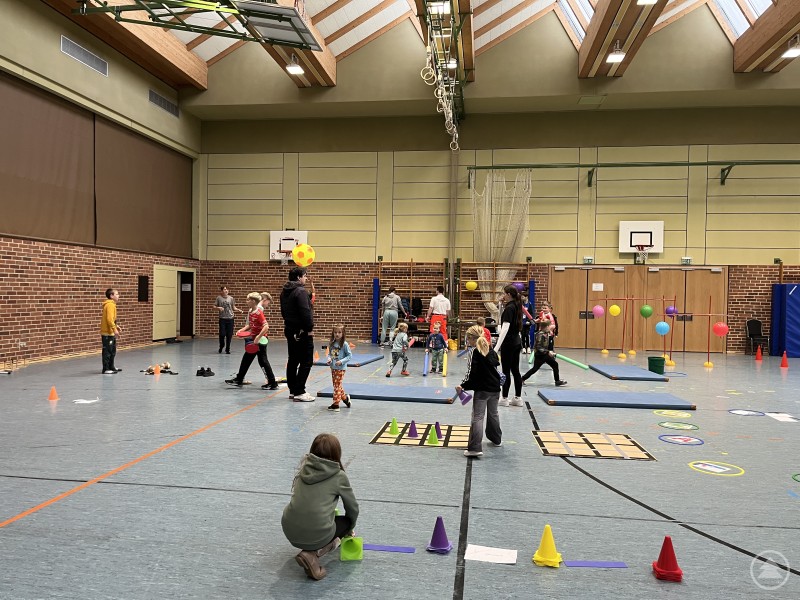 Kinder und Erwachsene bewegen sich in einer Turnhalle durch einen Parcours mit bunten Bällen, Hütchen und Spielgeräten.