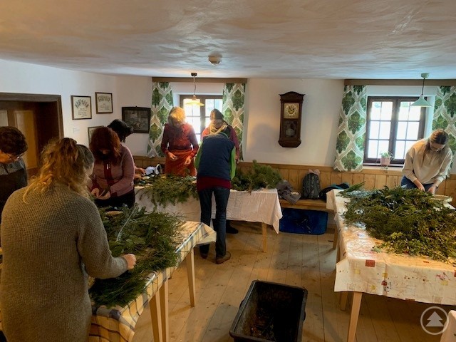 Mehrere Teilnehmerinnen stehen in einer gemütlichen Wirtsstube des Paul-Friedl-Hauses im Freilichtmuseum Finsterau und binden Adventskränze aus frischem Grün. Auf den Tischen liegen Zweige, Werkzeuge und Dekorationsmaterial.