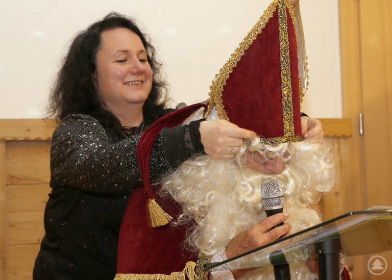 Eine Frau richtet dem als Nikolaus verkleideten Mann w&auml;hrend einer Weihnachtsfeier auf einer B&uuml;hne die Mitra, w&auml;hrend er ein Mikrofon in der Hand h&auml;lt.