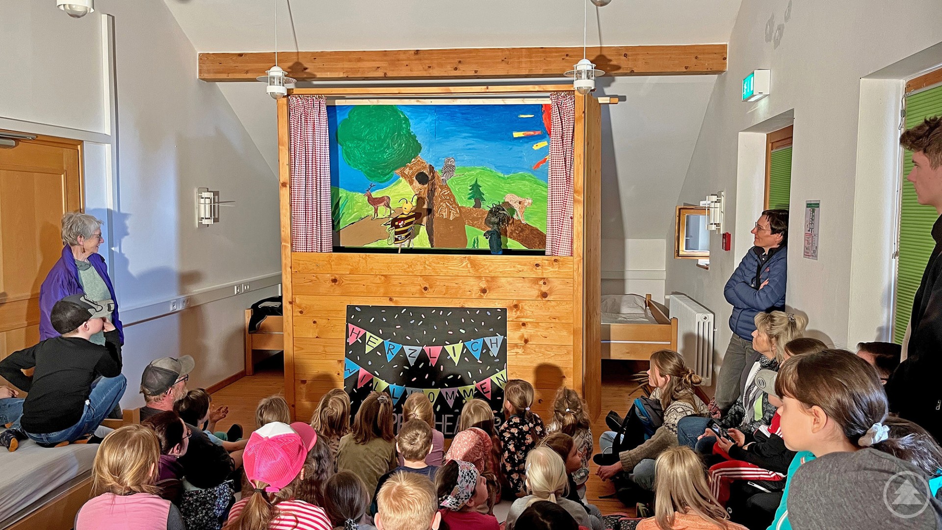 Vor vollbesetzten Rängen gaben Freiwillige des Nationalparks eine bejubelte Puppentheater-Vorstellung Das Bild zeigt eine Gruppe von Kindern und einigen Erwachsenen, die gespannt einem Puppentheaterstück zusehen. Die Bühne ist in ein hölzernes Podest mit Vorhängen integriert und zeigt eine liebevoll gemalte Waldszene mit Tieren. Über der Puppenbühne sind Figuren und eine Kulisse mit Naturmotiven (Bäume, Rehe, Vögel) zu sehen. Im Vordergrund sitzt das Publikum auf dem Boden, auf Bänken und Betten, während eine bunte „Herzlich Willkommen“-Tafel den Rahmen für die Veranstaltung bildet.