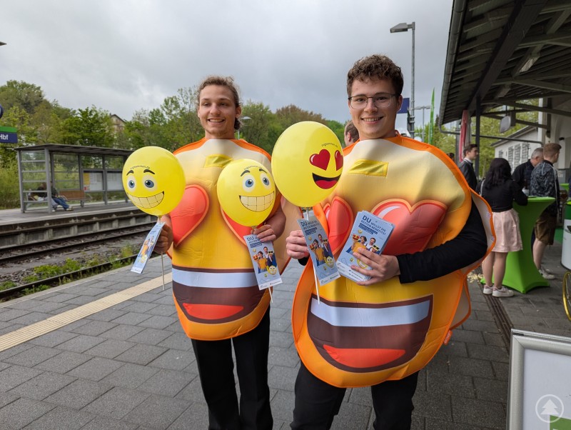 Weil gute Laune ansteckend ist - auch ohne strahlenden Sonnenschein Zwei als Emoji verkleidete Studierende verteilen Luftballons und Flyer