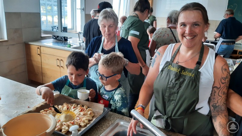 Teamgeist zeigten die Gäste in der Lehrküche. Der Nachwuchs war mit Eifer dabei! Eine Frau mit Schürze lächelt in die Kamera, während zwei Kinder neben ihr eine gebackene Pizzasonne dekorieren.