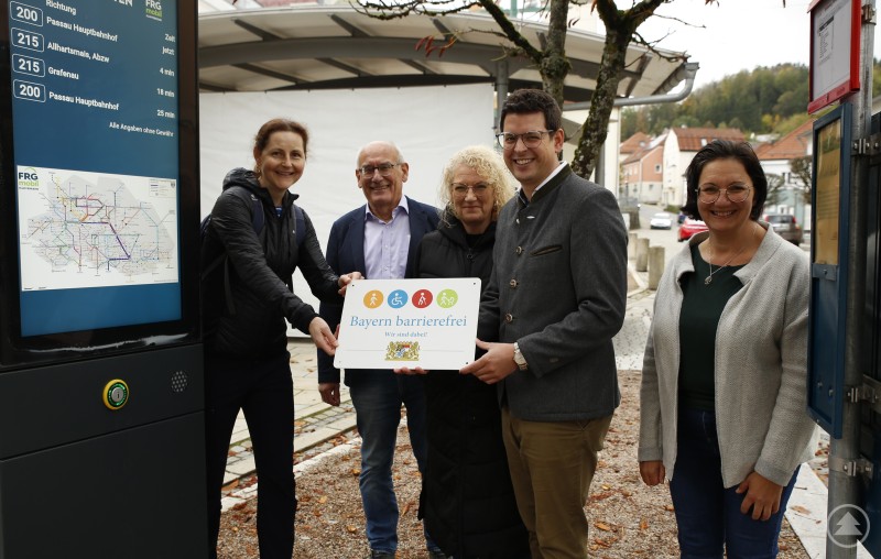 Im Auftrag der Bürgerinnen und Bürger mit Handicaps wie kurzen Beinen, Stock, Rollator, Rolli oder Einschränkungen bei Sinnen nahmen Yvonne Stadler (v.l.), Zweiter Bürgermeister Günter Klampfl, Doris Rötzer, Martin Pichler und Veronika Egger das Signet mit Piktogrammen für praktisch alle gerne entgegen. Foto: Markt Schönberg Fünf Personen stehen vor einem digitalen Fahrplananzeiger an einer Bushaltestelle in Schönberg. Sie halten gemeinsam ein Schild mit dem bayerischen Signet für Barrierefreiheit.