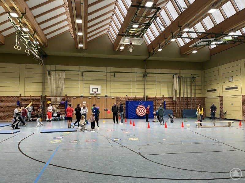 Geschicklichkeit gefragt – die Bewegungshalle war ein Publikumsmagnet für Jung und Alt. In einer Sporthalle nehmen Kinder und Erwachsene an Geschicklichkeits- und Wurfspielen teil, Zielscheiben und bunte Markierungen sind aufgestellt.