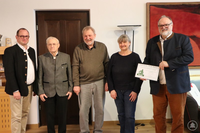 (Von rechts) Oberbürgermeister Jürgen Dupper, Renate Wögerbauer, Wolfgang Stiepani, Franz Nodes sowie Kulturreferent Dr. Bernhard Forster. Fünf Personen stehen nebeneinander in einem Innenraum, zwei Männer tragen traditionelle Jacken. Die Frau in der Mitte und der Mann rechts halten gemeinsam einen großen Gutschein mit dem Aufdruck „Stadt Passau“. Im Hintergrund sind Kunstwerke und Möbel zu sehen.