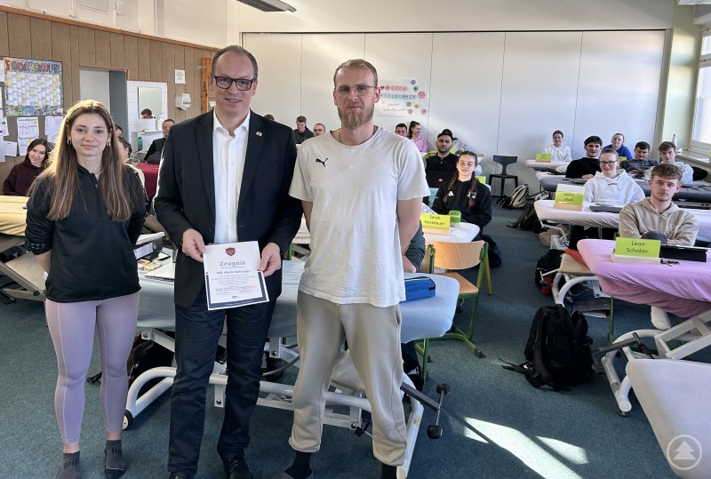 Als Dank für einen Vormittag direkten Sozialkundeunterricht gab es für MdL Martin Behringer ein Schulzeugnis mit Lob für besonders informatives Betragen von Jasmin Grübl und Tobias Tahedl als Klassensprecher. Ein Politiker steht in einem Klassenraum zusammen mit zwei Schülerinnen und Schülern vor einer Physiotherapieklasse und hält ein symbolisches Zeugnis in der Hand, während die Auszubildenden an Behandlungsliegen sitzen.
