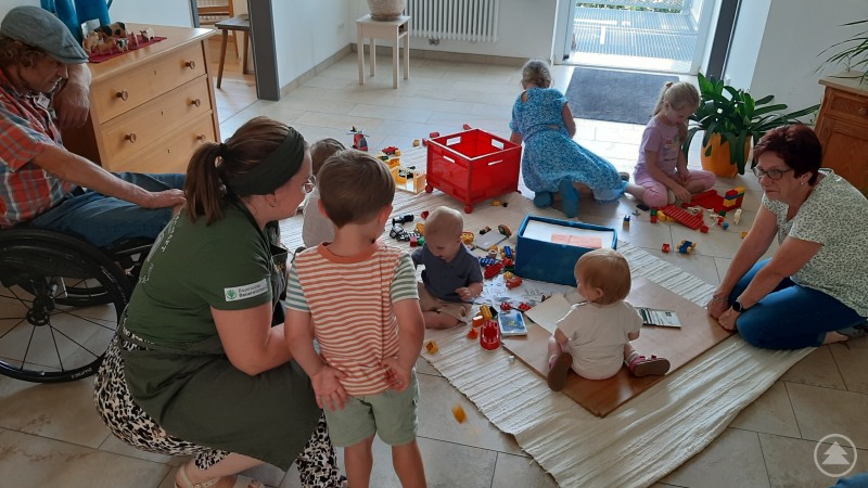 Beaufsichtigt von Eltern oder Großeltern schlossen die Kleinsten in der Spielecke neue Freundschaften. Mehrere kleine Kinder spielen auf einer Decke mit Spielzeug, Eltern oder Großeltern sitzen daneben.
