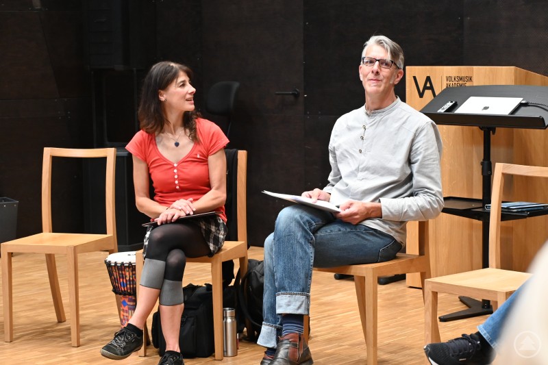 Die beiden Musiktherapeuten Gaby Flossmann und Stephan Maier zeigten den Anwesenden, wie Musik für Demenzbetroffene zu einem Schlüssel werden kann, der Erinnerungen weckt und ein Stück Lebensqualität zurückbringt. Eine Frau und ein Mann sitzen nebeneinander auf Stühlen in einem hellen Saal. Beide halten Unterlagen in den Händen, wirken konzentriert und im Austausch. Im Hintergrund steht ein Rednerpult mit der Aufschrift „Volksmusik Akademie“.