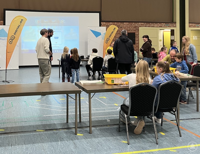 Kinder und Erwachsene sitzen an Tischen und arbeiten konzentriert, während vorne digitale Spiele mit einer großen Leinwand gezeigt werden.