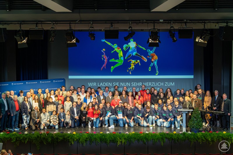 Sportlerehrung 2025 in Deggendorf Eine große Gruppe von Menschen steht auf einer Bühne in mehreren Reihen. Im Hintergrund ist eine große Leinwand mit sportlichen Silhouetten zu sehen, die zu einer Sportlerehrung einlädt.