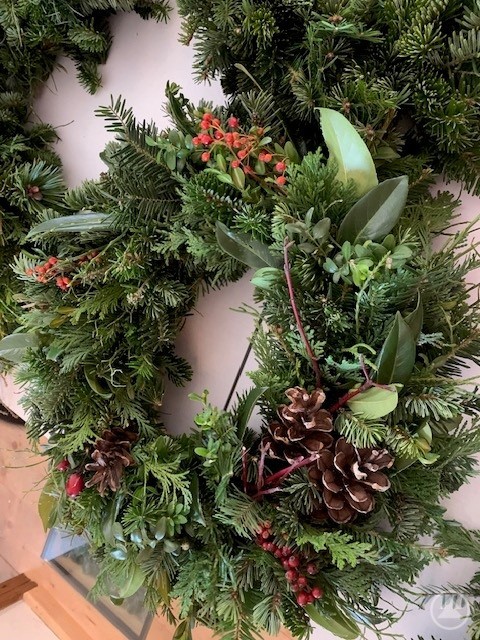 Nahaufnahme eines Adventskranzes aus verschiedenen Tannensorten, verziert mit Zapfen, roten Beeren und grünen Blättern.