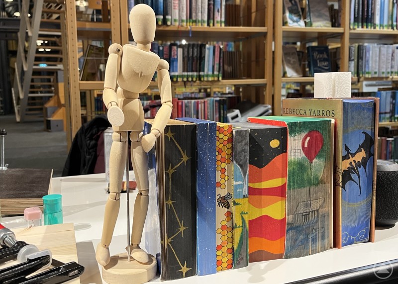 Carmen Knorrs bemalte Bücher sorgen für Inspiration Auf einem Tisch stehen verschiedene Bücher mit Farbschnitten. Ein Buch wurde dunkel bemalt mit Sternbildern, andere mit bunten Landschaften oder Symbolen. Daneben an den Büchern lehnt eine kleine Holzfigur mit flexiblen Gliedern.