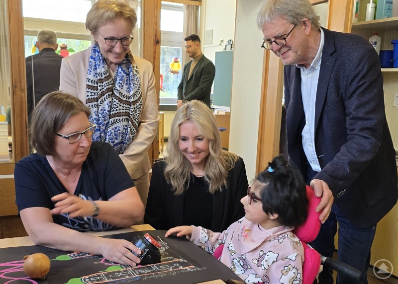Mehrere Erwachsene und ein Kind sitzen gemeinsam an einem Tisch in einem Klassenzimmer und besch&auml;ftigen sich mit Lernmaterialien. Eine Frau erkl&auml;rt etwas, w&auml;hrend die anderen aufmerksam zusehen.