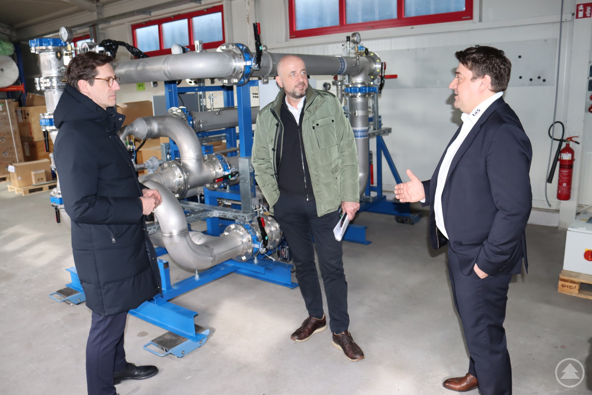 MdL Dr. Stefan Ebner (li.) lässt sich von den beiden UAS-Geschäftsführern Thomas Donaubauer und Martin Möginger (re.) die Funktionsweise einer Anlage zur optimierten und automatisierten Befeuerung für Glaswannen erläutern. Drei Männer stehen vor einer großen, komplexen technischen Anlage aus Edelstahlrohren in einer Werkhalle. Zwei der Männer tragen Anzug, einer eine grüne Jacke. Sie unterhalten sich angeregt.