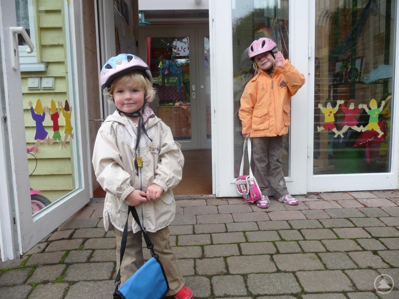 Gut vorbereitet für den Start: Kinder vor einer Einrichtung mit Schultasche und Helm, Symbol für den Übergang vom Kindergarten in die Schule. Zwei Kinder mit Fahrradhelmen stehen vor einem Eingang, vermutlich zu einem Kindergarten oder einer Schule. Beide tragen Jacken, kleine Taschen und wirken fröhlich. An den Glasscheiben sind bunte Kinderfiguren aus Papier angebracht.