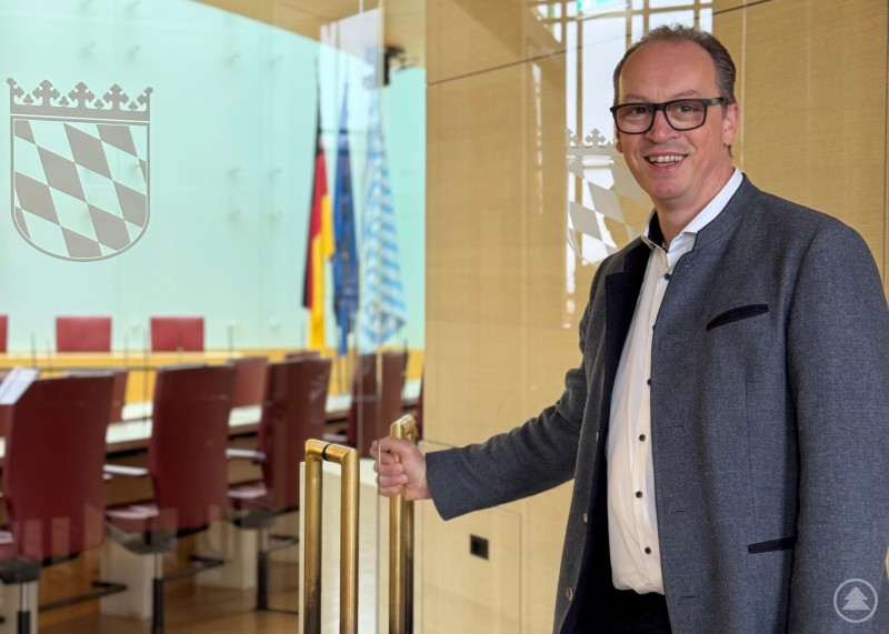 Gute Nachrichten aus dem Bayerischen Landtag: Martin Behringer freut sich über Millionen für Freyung. Martin Behringer öffnet eine Glastür zu einem modern gestalteten Sitzungssaal mit bayerischem Staatswappen und Flaggen im Hintergrund.