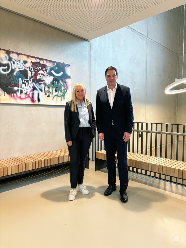Schulleiterin Elvira Wudy-Engleder und Landrat Sebastian Gruber am Staatlichen Beruflichen Schulzentrum Waldkirchen. Eine Frau und ein Mann stehen gemeinsam in einem modernen Flur mit Betonelementen, Holzbänken und Graffiti-Kunst an der Wand. Beide tragen Business-Outfits und lächeln in die Kamera.