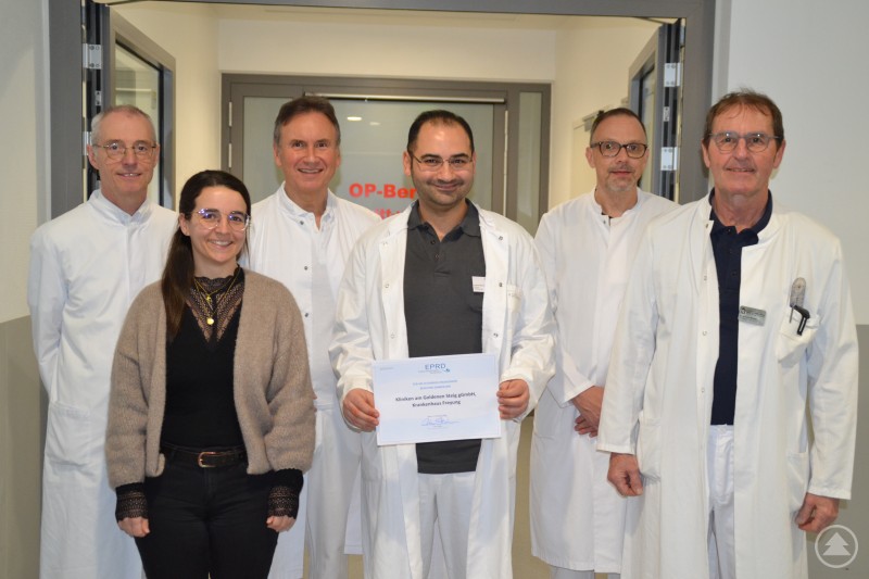 Ärzteteam der Kliniken Am Goldenen Steig mit dem EPRD-Zertifikat für über zehn Jahre Teilnahme am Endoprothesenregister Deutschland. Mehrere Ärzte und eine Mitarbeiterin stehen im Krankenhausflur vor dem OP-Bereich. In der Mitte hält ein Arzt ein Zertifikat des Endoprothesenregisters Deutschland, während das Team der Kliniken Am Goldenen Steig gemeinsam für ein Gruppenfoto posiert.