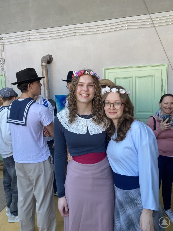 Zwei Sch&uuml;lerinnen in historischen Kleidern mit Blumenkr&auml;nzen stehen l&auml;chelnd vor einer T&uuml;r im Atelier und posieren f&uuml;r ein Foto.