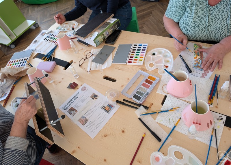 Vier Kursteilnehmer sitzen an einem Tisch der mit Pinseln, Schablonen, B&uuml;chern und Stiften &uuml;bers&auml;ht ist und erstellen ganz individuelle Entw&uuml;rfe zu ihren B&uuml;chern.