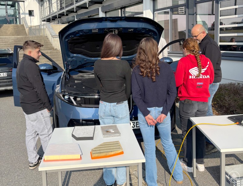 Der Blick unter die Motorhaube. Mechatronik gehörte zu den Themen, die die Schülerinnen und Schüler besonders interessierten. Mehrere Schülerinnen und Schüler stehen um ein Auto mit geöffneter Motorhaube und lassen sich technische Details erklären.