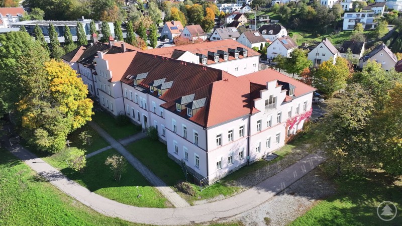 Das ehemalige Elisabethenheim in Deggendorf – hier soll künftig eine Kindertageseinrichtung entstehen. Luftaufnahme eines großen historischen Gebäudes mit hellrosa Fassade und roten Dächern, umgeben von Bäumen mit herbstlicher Färbung und einem Fußweg im Vordergrund. Im Hintergrund sind Wohnhäuser und begrünte Hänge zu sehen.