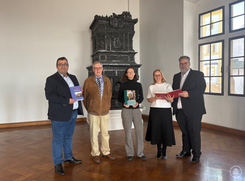 (v.l.n.r.) Dr. Markus Eberhardt, Dr. Franz-Reiner Erkens, Dr. Teresa Lohr, Dr. Stefanie Buchhold und Bürgermeister Andreas Rother präsentieren die neue Publikation zu Ferdinand Wagner im Oberhausmuseum Passau. Fünf Personen stehen in einem hellen Museumsraum mit altem, reich verziertem schwarzen Kachelofen. Alle halten Bücher in den Händen und blicken freundlich in die Kamera.
