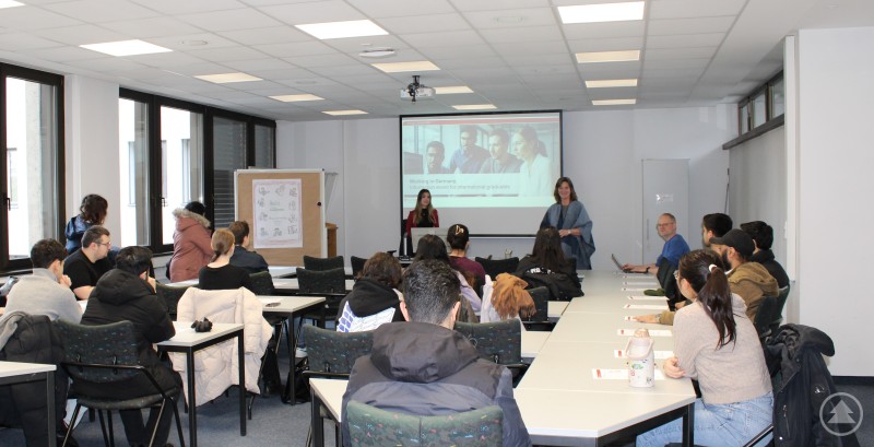 Bildunterschrift vorne von links: Tanja Grauberger (Berufsberaterin im Erwerbsleben), Christine Reitberger (Teamleiterin Berufsberatung vor dem Erwerbsleben), Benedikt Oswald (Berufsberater vor dem Erwerbsleben) In einem Seminarraum der Agentur für Arbeit Passau sitzen internationale Studierende an Tischen und hören einen englischsprachigen Vortrag, vorne ist eine Präsentation zu sehen.