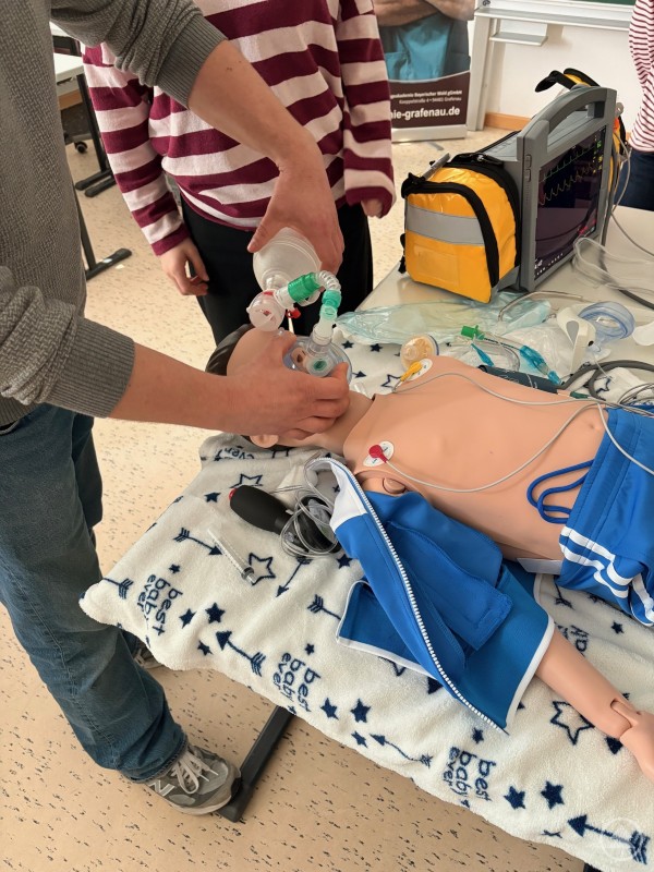 Auch der Bereich der Pflegeberufe war auf dem Handwerkstag vertreten. Am Stand der Pflegeakademie Bayerischer Wald durften die Schülerinnen und Schüler Vitalfunktionen prüfen und Zugänge legen. An einem Tisch wird an einer Übungspuppe medizinisches Equipment demonstriert, während Schülerinnen und Schüler zuschauen.