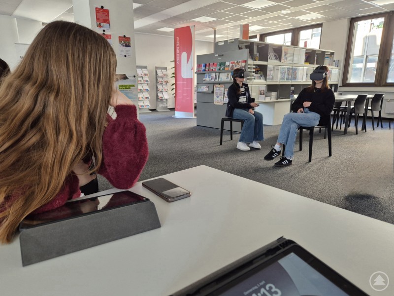 Das Bild zeigt eine Szene in einem modernen, hellen Raum – vermutlich eine Bibliothek oder ein Lernzentrum. Im Vordergrund sitzt eine Person mit langen Haaren an einem Tisch, auf dem ein Tablet und ein Smartphone liegen. Im Hintergrund sitzen zwei weitere Personen auf Stühlen, die Virtual-Reality-Brillen tragen. Sie scheinen in eine VR-Anwendung vertieft zu sein. Regale mit Informationsmaterialien und Prospekten sowie Plakate sind ebenfalls zu sehen. Die Szene deutet auf eine Bildungseinrichtung oder ein Projekt mit digitalen Lernmethoden hin.