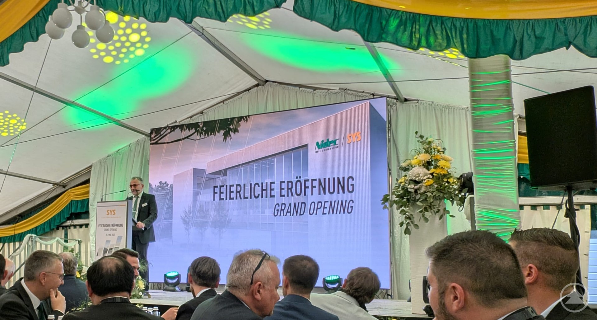 Geschäftsführer Werner Borth begrüßt die Gäste bei der feierlichen Eröffnung des neuen Nidec SYS Gebäudes in Grafenau. Mann im Anzug spricht am Rednerpult auf einer Bühne vor einem großen LED-Screen mit der Aufschrift „Feierliche Eröffnung – Grand Opening“ im Festzelt.