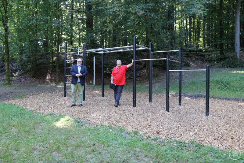 Oberbürgermeister Jürgen Dupper (links) besichtigt den Motorikpark Ingling zusammen mit Franz Kessel (rechts), Dienststellenleiter Schul- und Sportamt. Zwei Männer stehen vor einer neuen Edelstahl-Kletter- und Trainingsanlage im Freien. Die Anlage ist mit Rindenmulch umgeben und liegt in einem bewaldeten Bereich. Einer der Männer trägt einen roten Pullover, der andere ein dunkles Sakko.