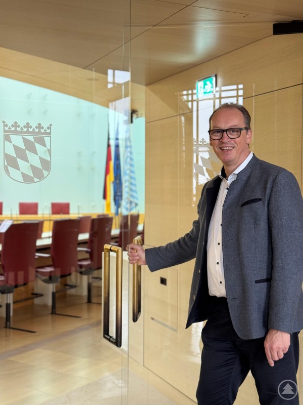 MdL Martin Behringer kann den Besuch des Maximilianeums nur empfehlen. Hier begegnen sich Kultur, Geschichte und Politik in besonderer Weise. Wer im Herbst samstags oder sonntags nach München reist, kann einen faszinierenden Abstecher jetzt schon anmelden. Ein Mann in Trachtenjacke steht an einer Glastür mit dem bayerischen Wappen und öffnet den Eingang zu einem Sitzungssaal mit roten Stühlen und Landesflaggen im Hintergrund.